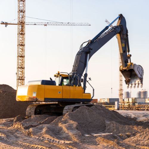 Excavadora amarilla moviendo arena en obra con grúas torre al fondo
